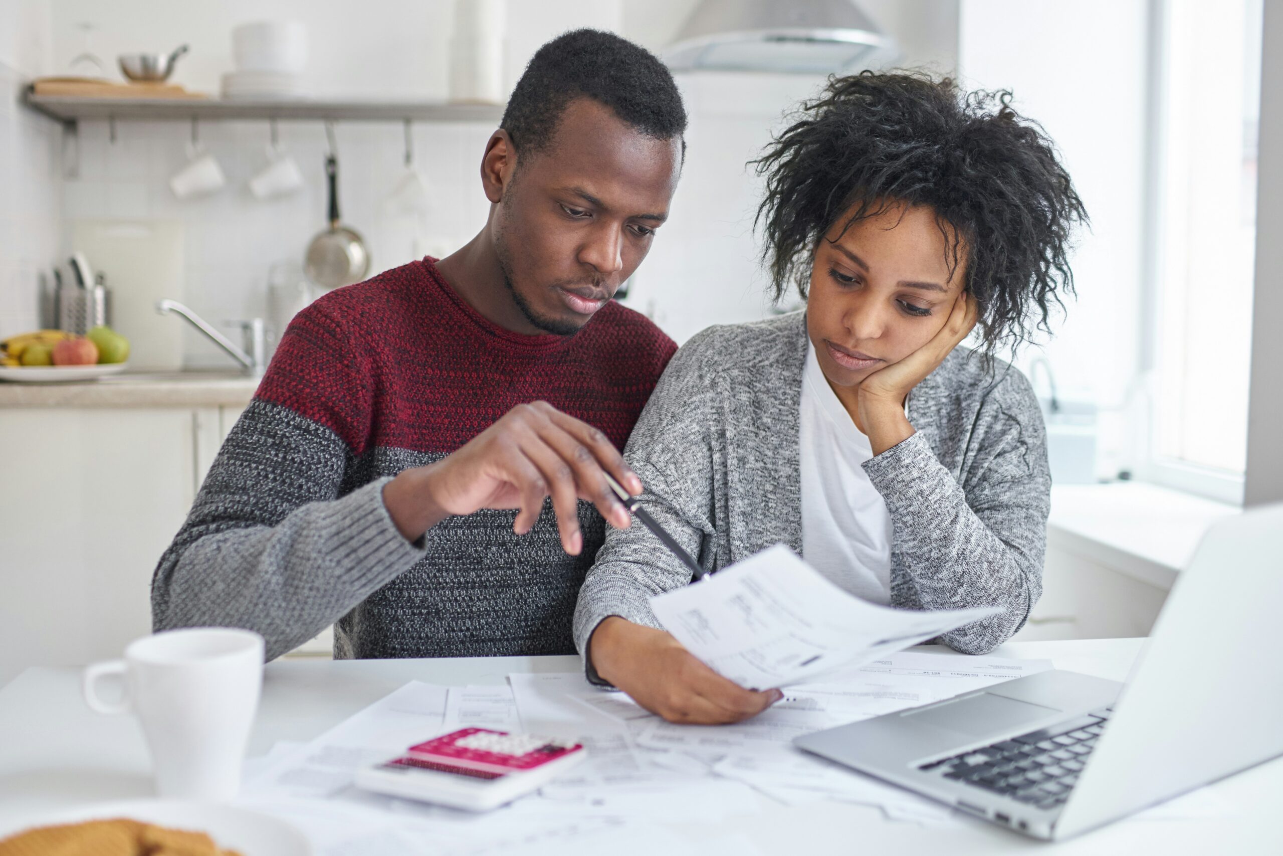 Couple reviewing household bills and financial documents together at a kitchen table with a laptop and calculator.